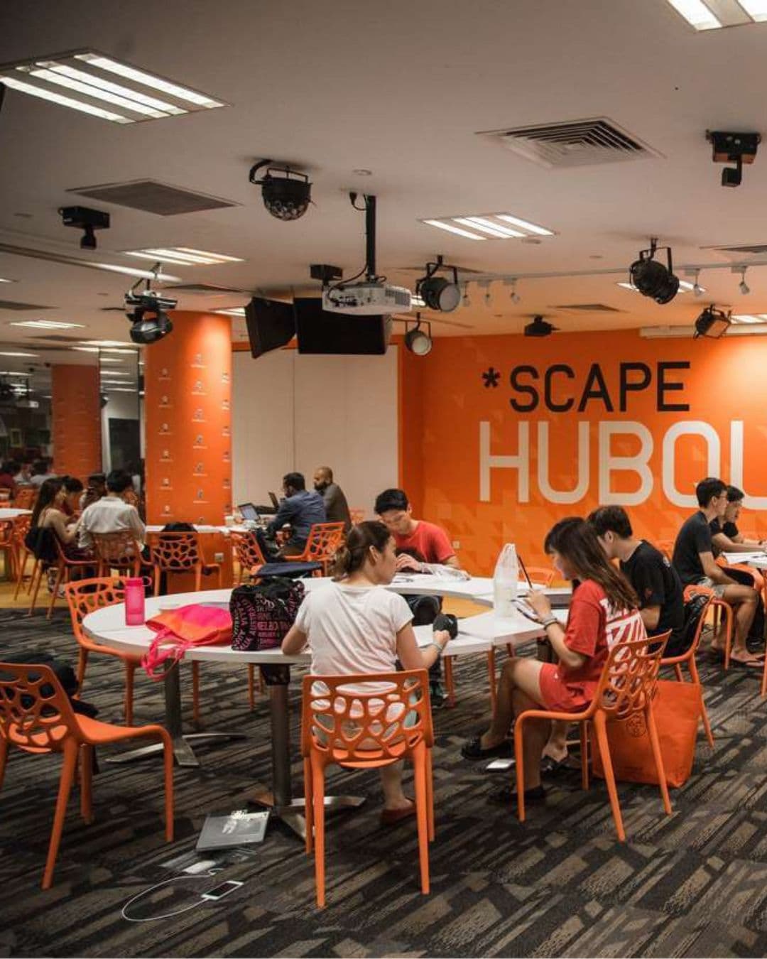 *SCAPE HubQuarters – Creative Collaborative Study Space Orange-and-white themed round desks with charging ports at *SCAPE HubQuarters